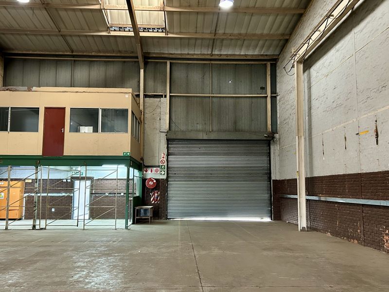 Spacious industrial warehouse featuring a high-clearance roller shutter door, concrete flooring, exposed steel roof structure, and an integrated mezzanine office level.