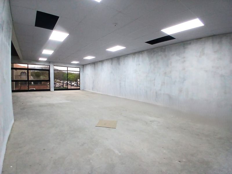 An empty, open-plan office space featuring a suspended ceiling with recessed lighting, large windows, and unfinished wall and floor surfaces.