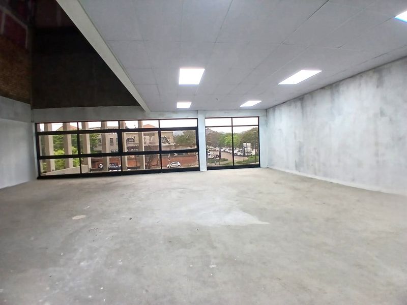 A spacious, empty office unit featuring large windows, a suspended ceiling with recessed lighting, and unfinished wall and floor surfaces.