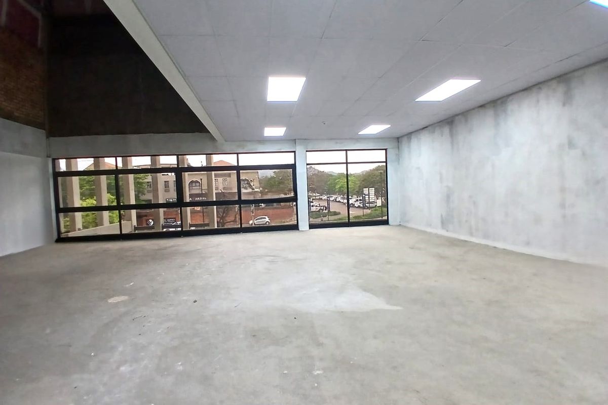 A spacious, empty office unit featuring large windows, a suspended ceiling with recessed lighting, and unfinished wall and floor surfaces.