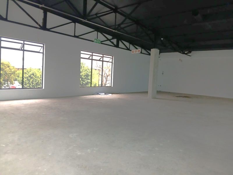 A spacious, open-plan retail unit featuring high exposed steel roof trusses, large windows for natural light, and a clean, unfinished concrete floor.