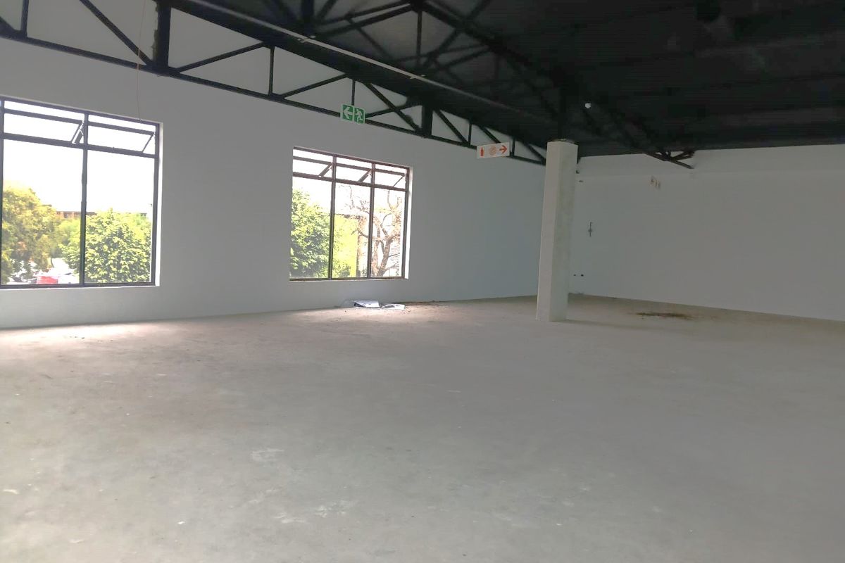 A spacious, open-plan retail unit featuring high exposed steel roof trusses, large windows for natural light, and a clean, unfinished concrete floor.