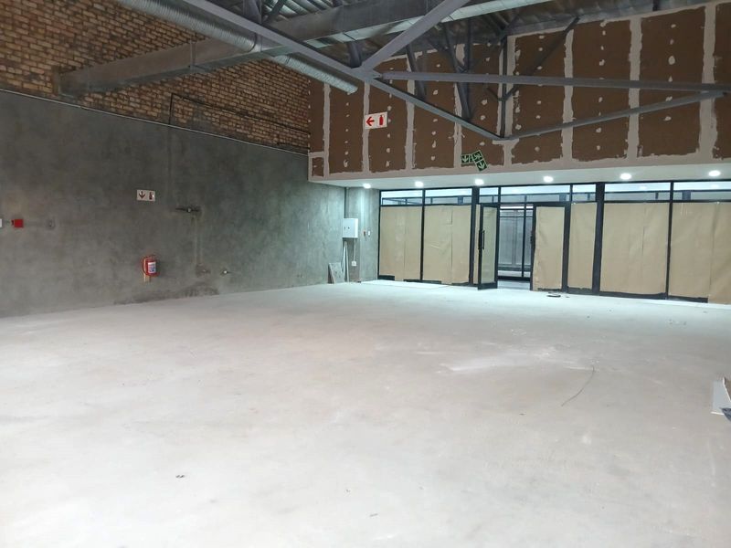 An expansive, open-plan retail space in shell condition, featuring exposed brick walls, steel roof trusses, and large glass storefront windows.