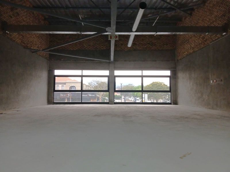 An expansive, unfinished open-plan retail space featuring exposed brickwork, industrial steel roof trusses, and large windows offering natural light and street views.