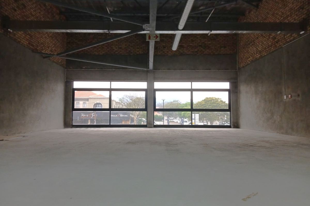 An expansive, unfinished open-plan retail space featuring exposed brickwork, industrial steel roof trusses, and large windows offering natural light and street views.