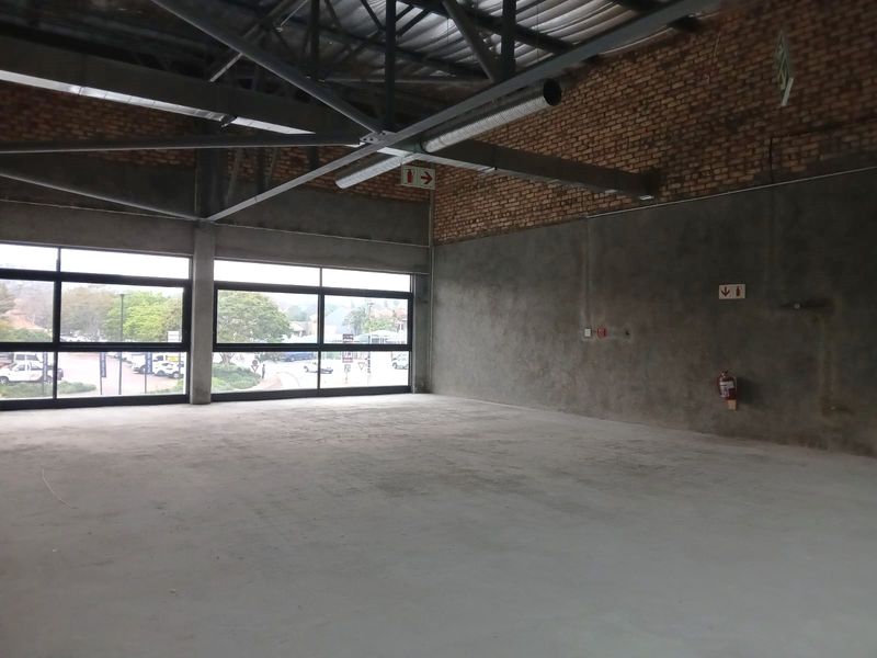 An open-plan industrial retail space featuring exposed brick walls, concrete flooring, high steel roof trusses, and large windows providing ample natural light.