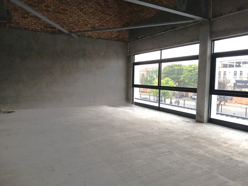 An expansive, unfinished open-plan retail space featuring exposed brickwork, concrete flooring, and large windows providing ample natural light.