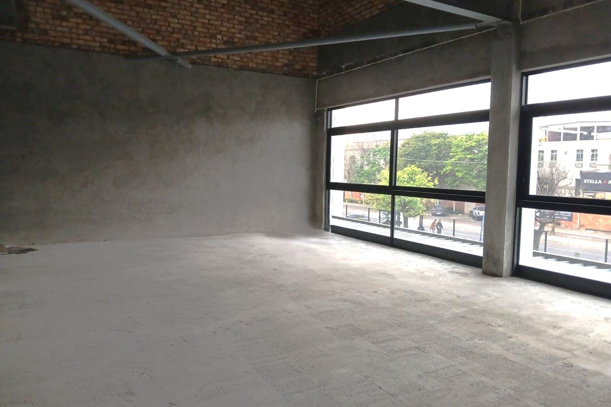 An expansive, unfinished open-plan retail space featuring exposed brickwork, concrete flooring, and large windows providing ample natural light.
