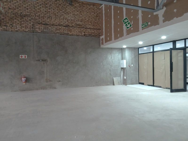 An unfinished retail utility space featuring exposed brickwork, concrete flooring, recessed lighting, and aluminium-framed glass entrance doors.
