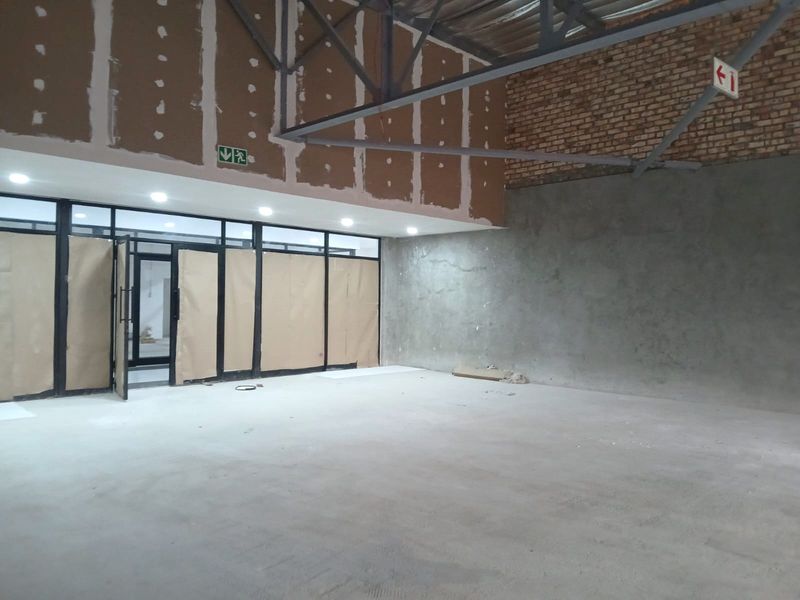 An unfinished open-plan retail space featuring exposed steel roof trusses, brickwork, and large aluminium-framed glass storefront windows, currently in a shell condition.