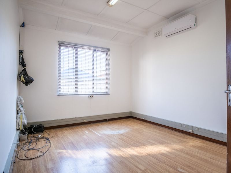 A clean, empty office space featuring laminate flooring, a wall-mounted air conditioning unit, and large windows with fitted venetian blinds.