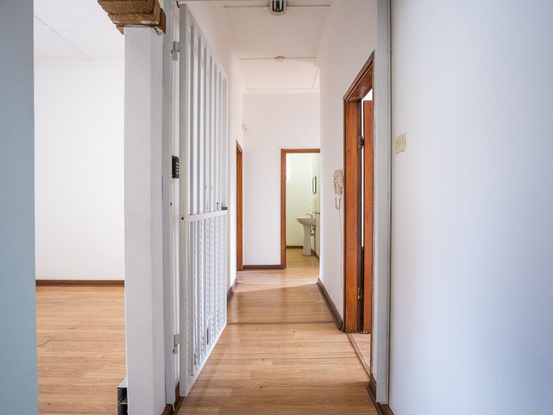A bright, empty office hallway featuring laminate flooring, wooden door frames, and a security gate, leading towards a secondary room with a basin.