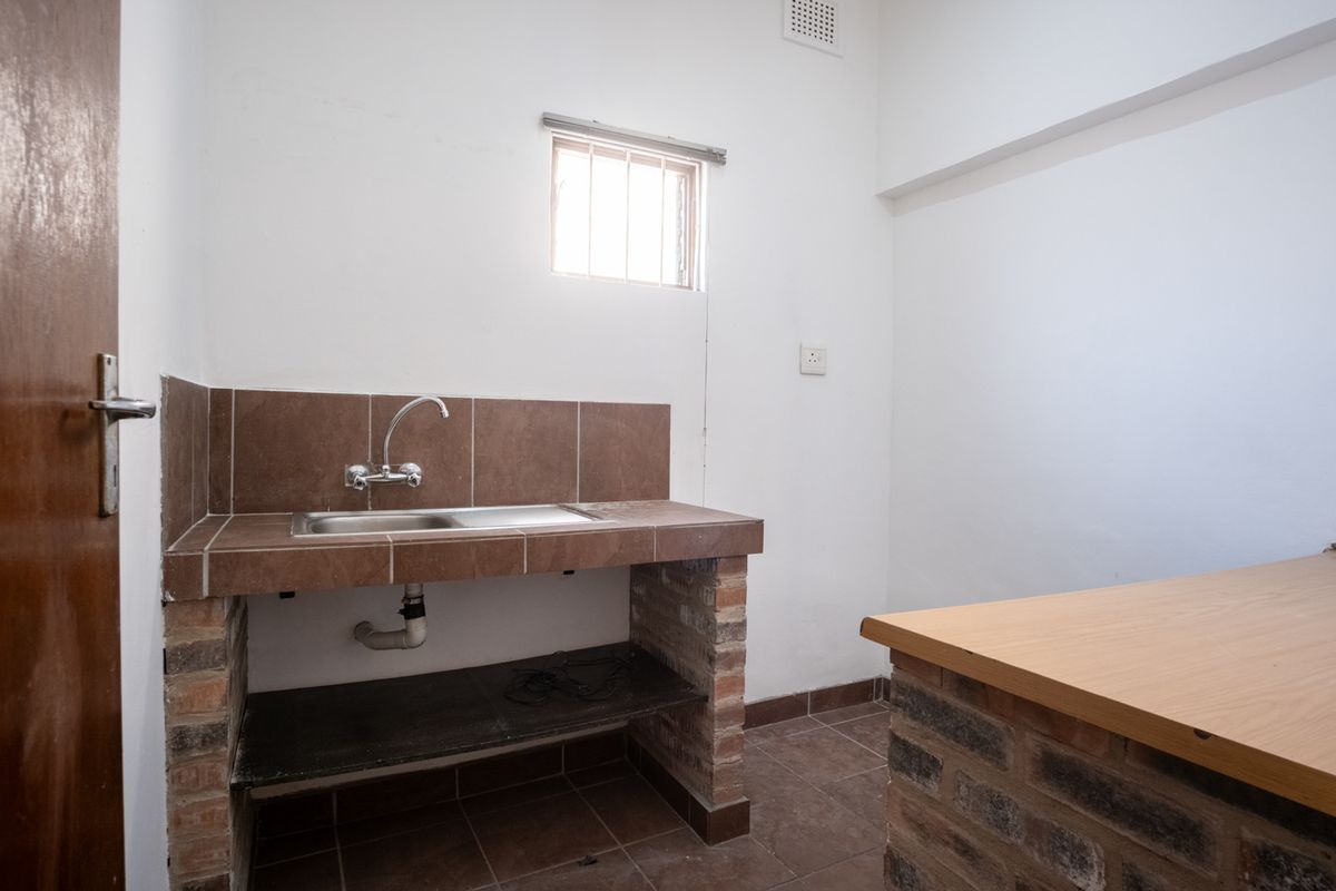 A basic kitchenette featuring a tiled counter with a stainless steel sink, brick base construction, and a small window with security bars.