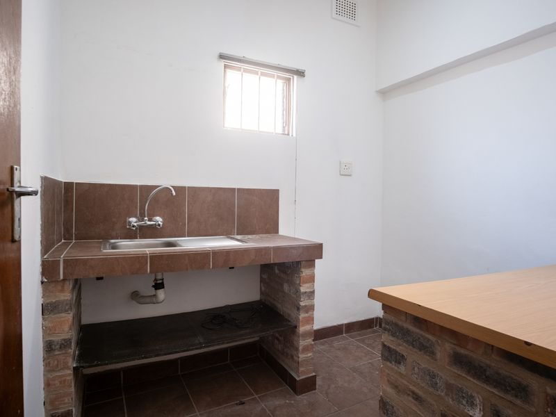 A basic kitchenette featuring a tiled counter with a stainless steel sink, brick base construction, and a small window with security bars.