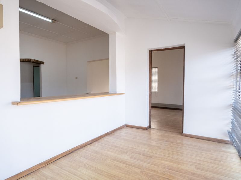 Empty office space featuring laminate flooring, a serving hatch, and timber window blinds, offering a versatile layout for professional use.