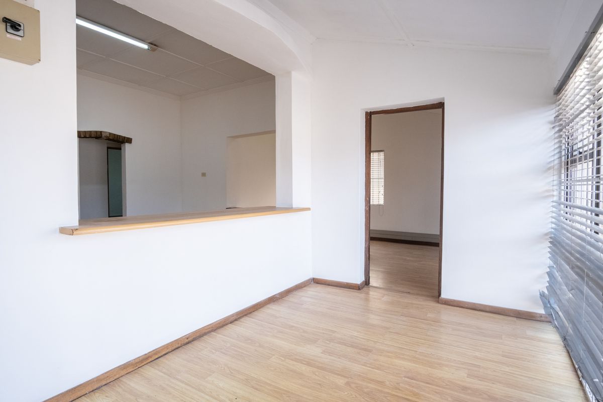 Empty office space featuring laminate flooring, a serving hatch, and timber window blinds, offering a versatile layout for professional use.