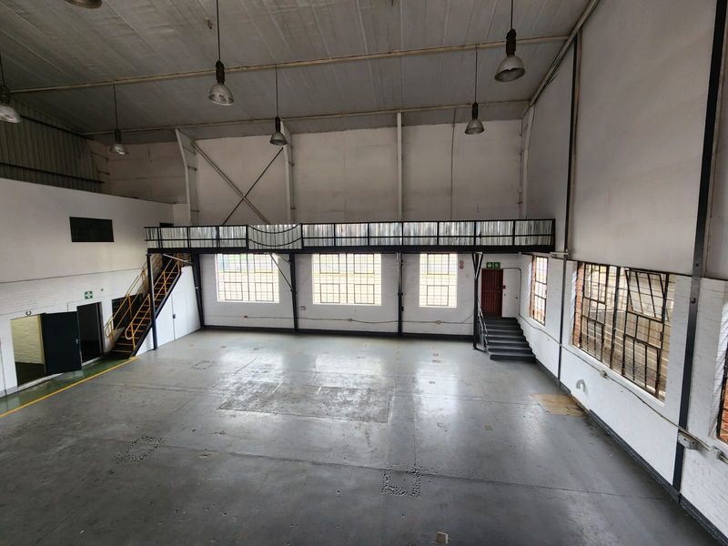 Spacious industrial warehouse unit featuring a mezzanine level, high ceilings with pendant lighting, steel-framed windows, and durable concrete flooring.