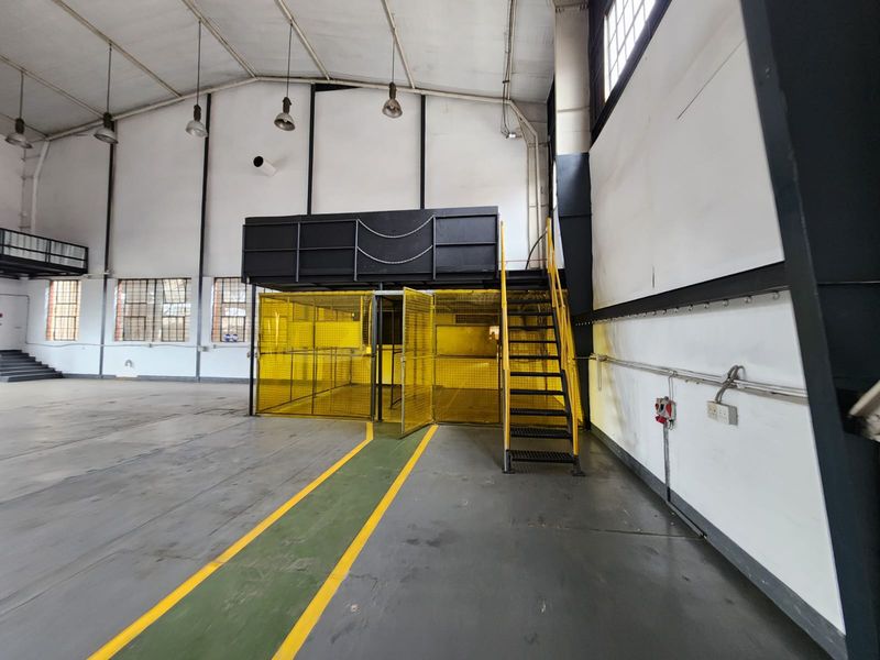 Spacious industrial utility area featuring a mezzanine level, metal staircase, and secure safety cage enclosure with high ceilings and concrete flooring.