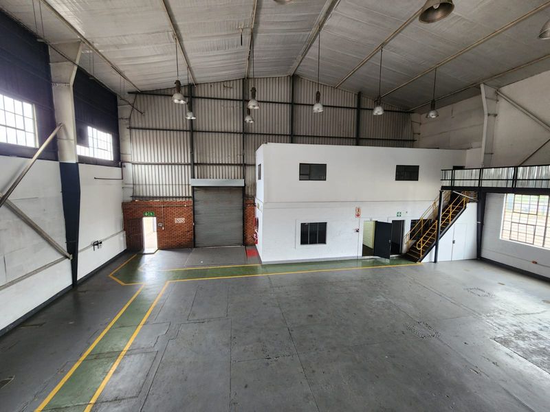 Spacious industrial warehouse featuring high ceilings, exposed steel trusses, a mezzanine level with staircase, roller shutter door access, and durable concrete flooring.