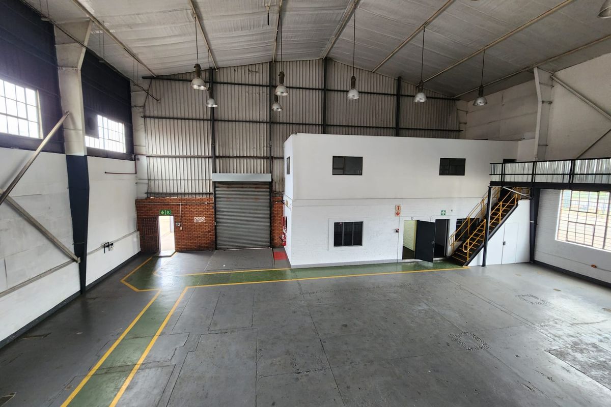 Spacious industrial warehouse featuring high ceilings, exposed steel trusses, a mezzanine level with staircase, roller shutter door access, and durable concrete flooring.