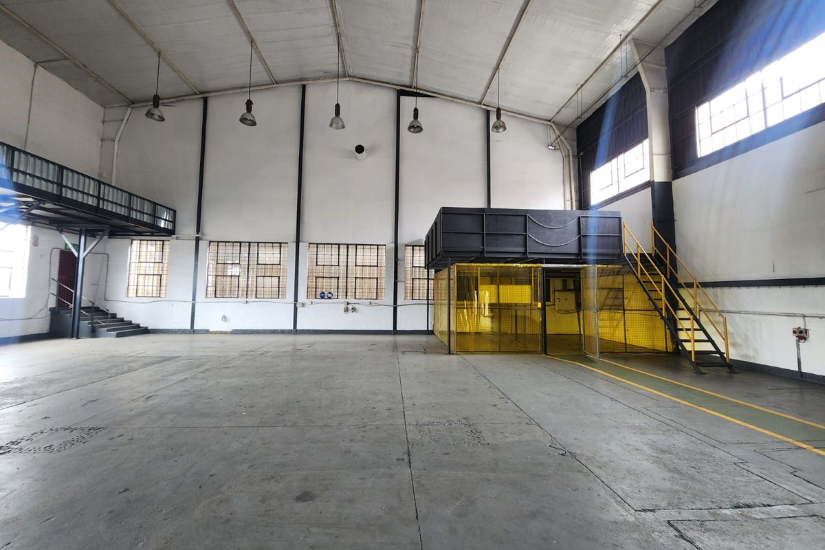 Spacious industrial warehouse area featuring high ceilings, concrete flooring, a mezzanine level with steel staircase, and secure partitioned workspace.