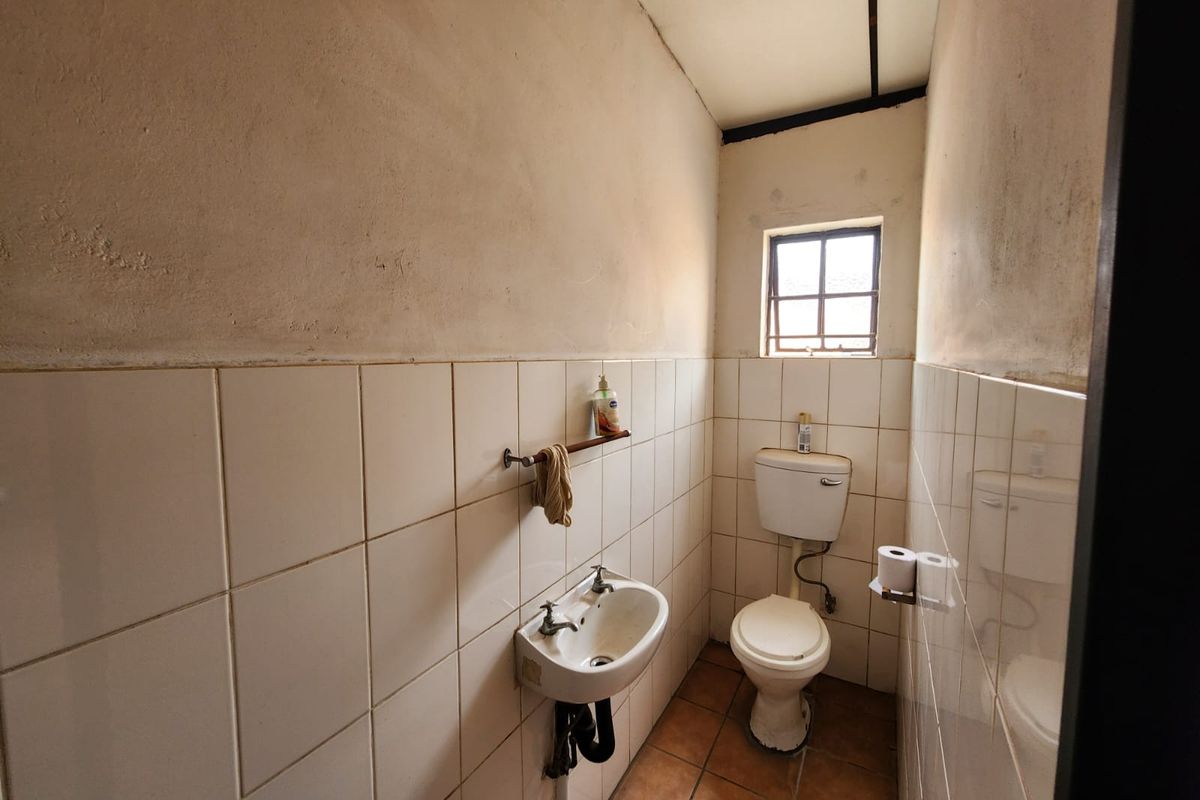 A compact industrial-style bathroom featuring a wall-mounted basin, ceramic toilet, and tiled finishes, requiring cosmetic updates to improve its overall condition.