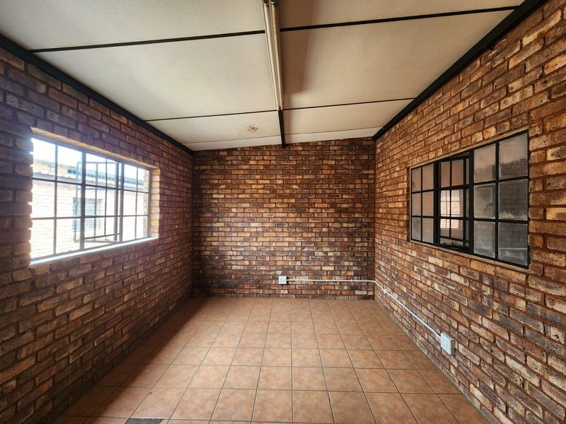 An empty industrial-style office space featuring exposed brick walls, tiled flooring, and steel-framed windows with surface-mounted electrical infrastructure.