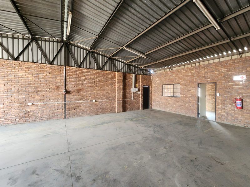 Spacious industrial utility area featuring exposed brick walls, high steel truss ceilings, concrete flooring, and functional lighting, suitable for storage or workshop use.