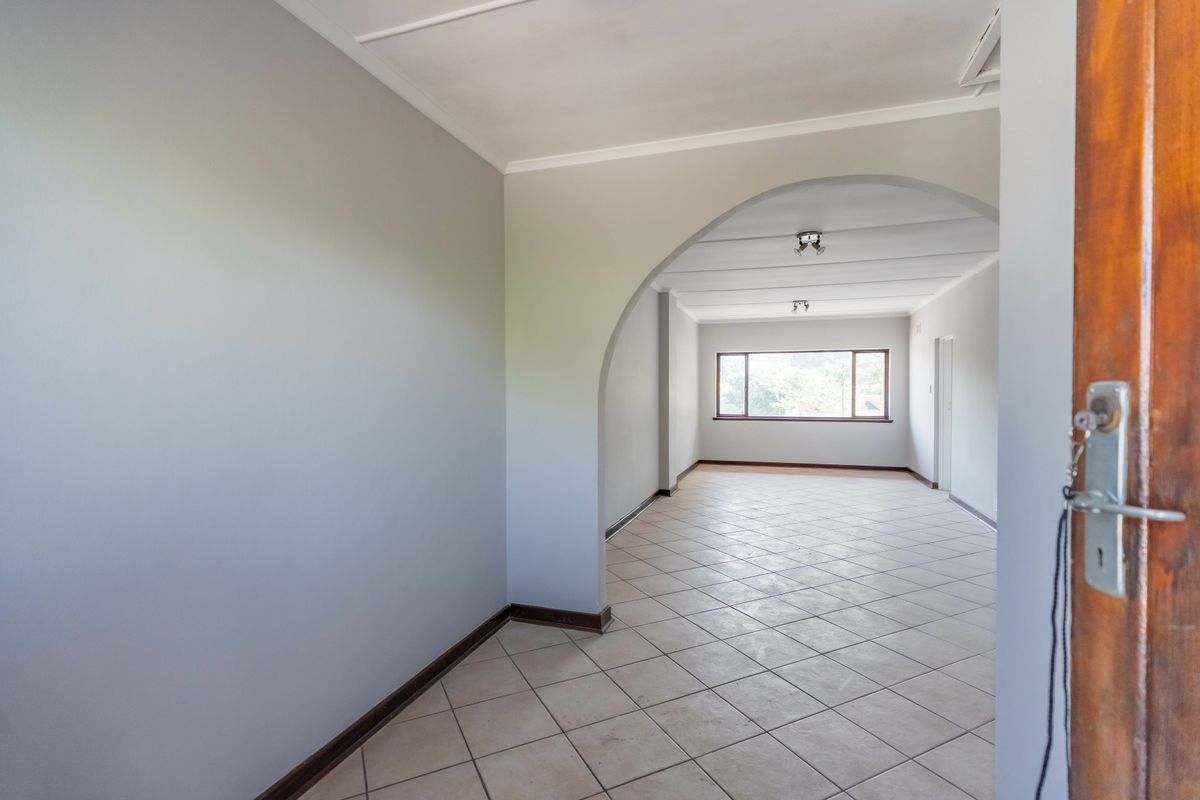 An empty, open-plan living area featuring neutral walls, tiled flooring, and a distinctive architectural archway leading to a bright, naturally lit space.