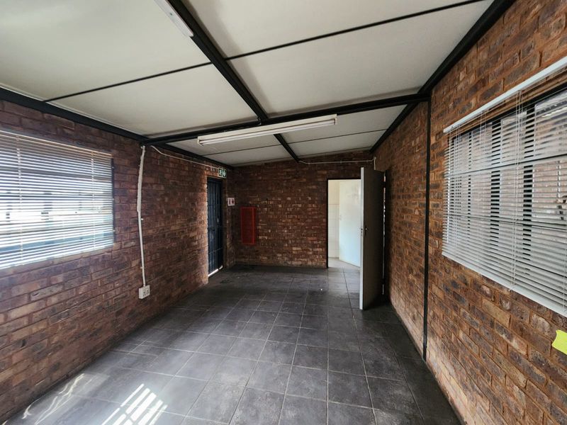 An empty industrial-style office space featuring exposed brick walls, tiled flooring, and functional fluorescent lighting with venetian blinds on the windows.