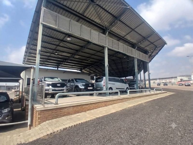 A commercial retail display area featuring a large steel-roofed canopy, brick retaining wall, and paved surface suitable for vehicle or product showcasing.