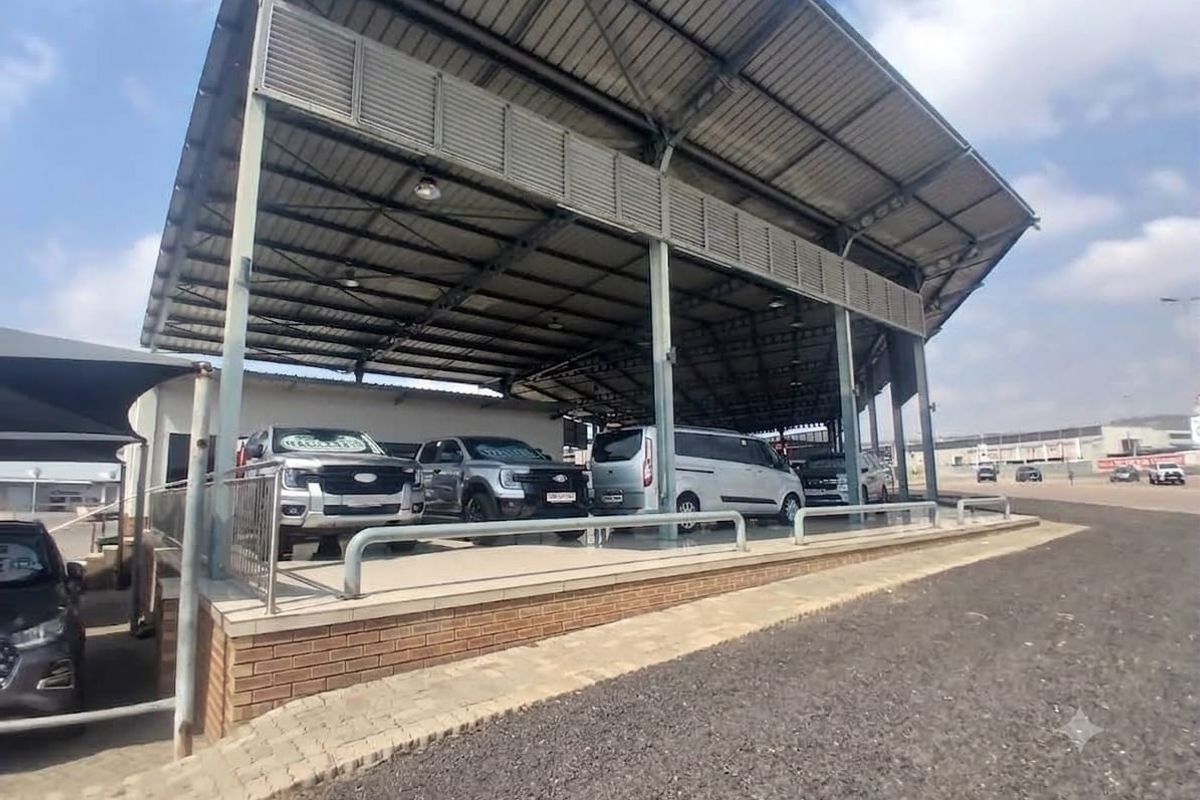 A commercial retail display area featuring a large steel-roofed canopy, brick retaining wall, and paved surface suitable for vehicle or product showcasing.