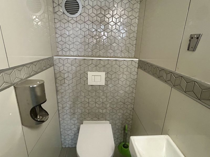A compact, modern bathroom featuring tiled walls with geometric accents, a wall-mounted toilet with a flush plate, and a small wall-mounted basin.