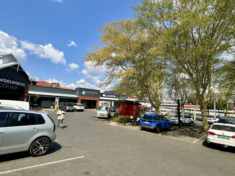 A view of the Eastwood Village retail center parking area, featuring established trees and prominent commercial signage for Woolworths and Clicks.