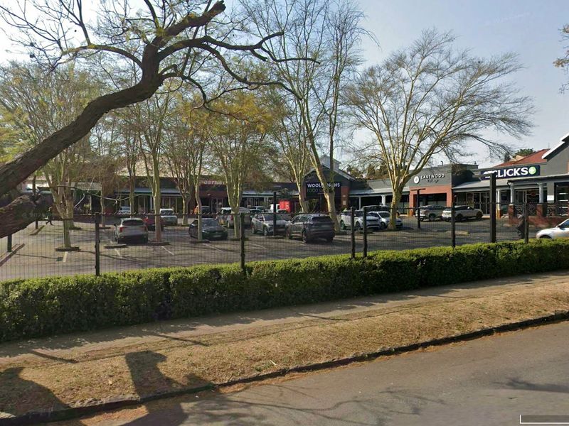 Street-level view of a retail shopping center featuring multiple storefronts, a paved parking lot, and perimeter fencing.