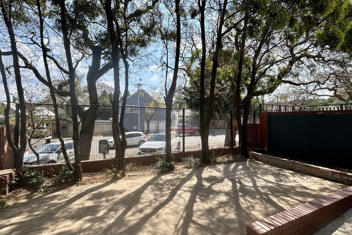 A paved outdoor area featuring brick retaining walls and mature trees, enclosed by metal security fencing with a view toward the street.