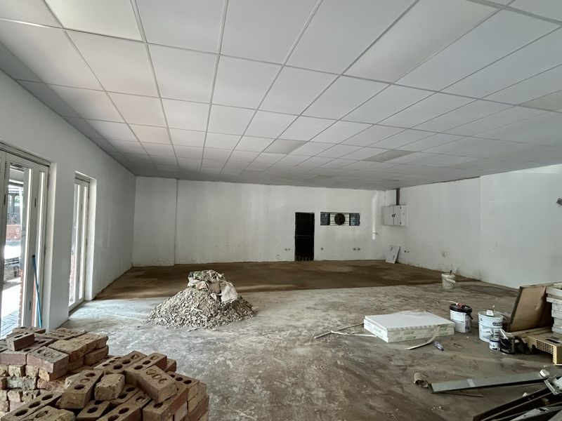 An open-plan retail space currently under renovation, featuring a suspended ceiling, large glass sliding doors, and unfinished concrete flooring.