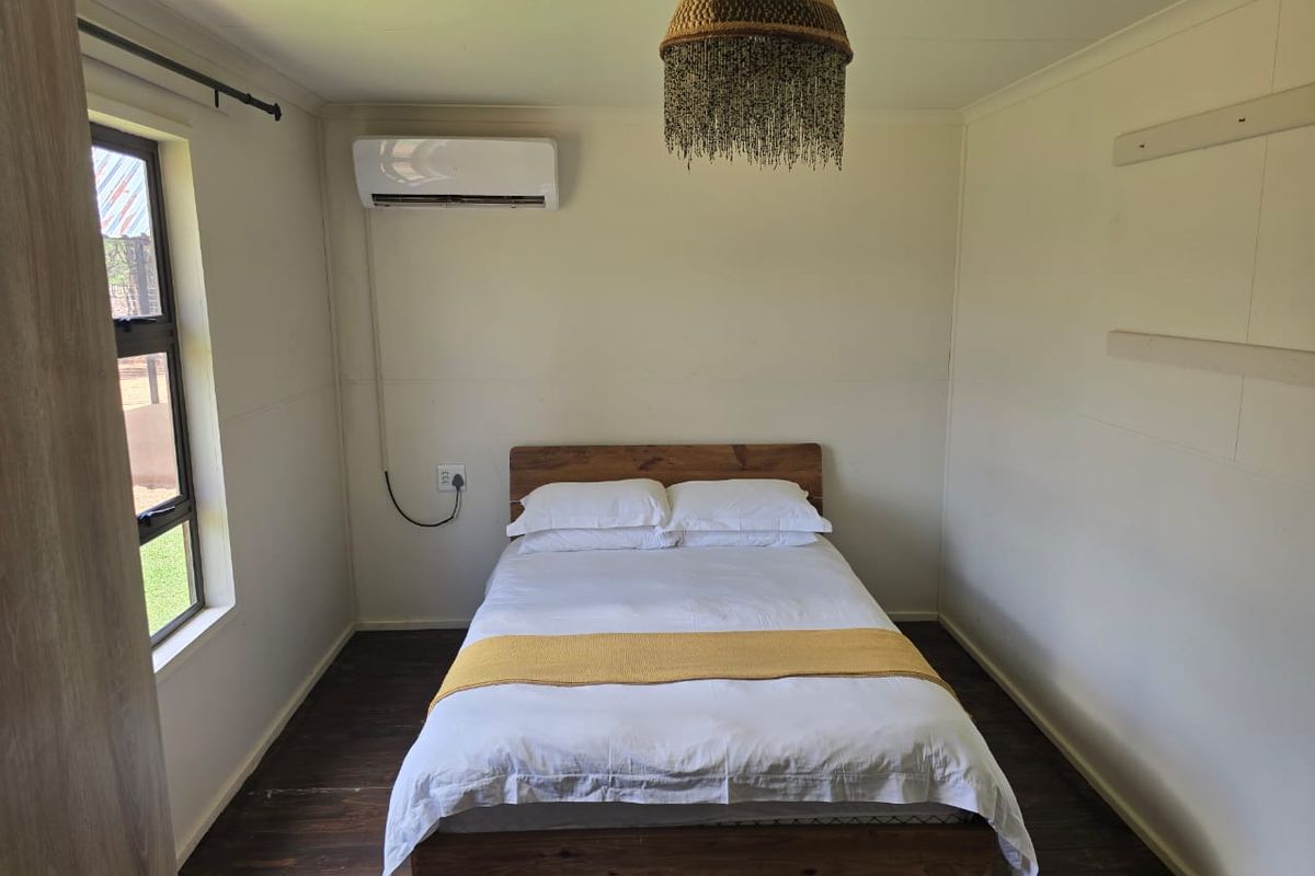 A simple bedroom featuring dark wood-look flooring, a wall-mounted air conditioning unit, and a window providing natural light.