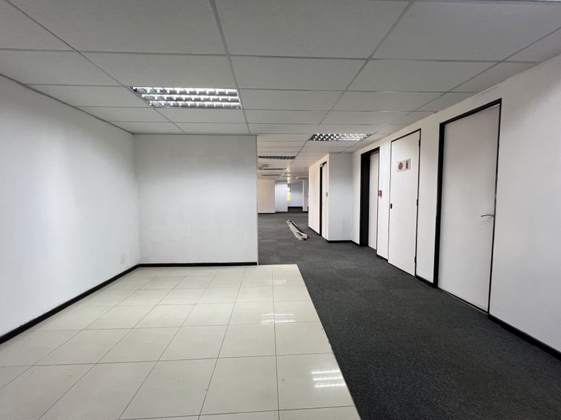 An empty, open-plan office space featuring a mix of tiled and carpeted flooring, suspended ceilings with recessed lighting, and multiple internal access doors.