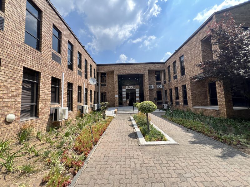 A well-maintained office building exterior featuring a face brick facade, paved central walkway, and landscaped garden beds leading to the main entrance.