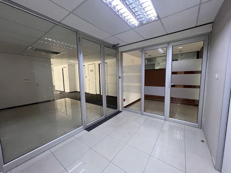 A bright, vacant office space featuring modern glass partitioning, aluminum frames, suspended ceilings with recessed lighting, and clean tiled flooring throughout.