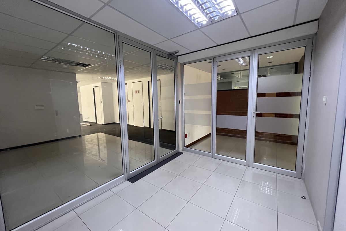 A bright, vacant office space featuring modern glass partitioning, aluminum frames, suspended ceilings with recessed lighting, and clean tiled flooring throughout.