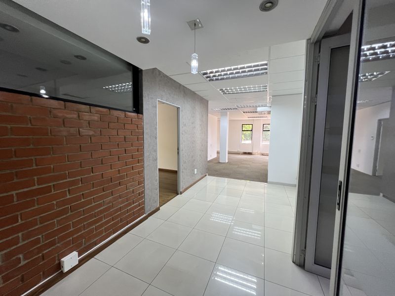 Modern office space featuring a combination of tiled and carpeted flooring, exposed brick accents, and recessed lighting throughout the open-plan layout.