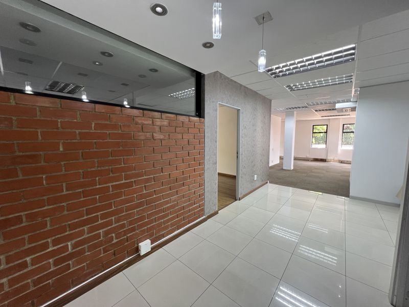 A bright, modern office space featuring a combination of tiled and carpeted flooring, exposed brick accents, and integrated ceiling lighting.