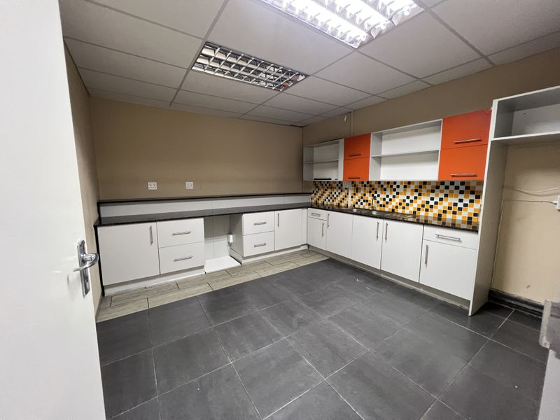 A clean, functional office kitchenette featuring white and orange cabinetry, a mosaic tile backsplash, dark floor tiles, and integrated lighting.