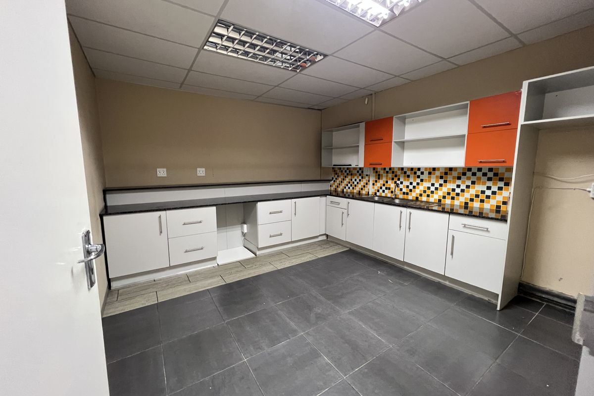 A clean, functional office kitchenette featuring white and orange cabinetry, a mosaic tile backsplash, dark floor tiles, and integrated lighting.