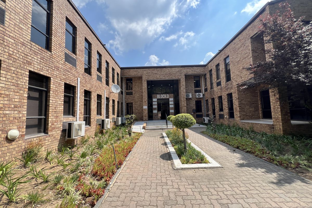A well-maintained office building exterior featuring a face brick facade, paved central walkway, and landscaped garden beds leading to the main entrance.