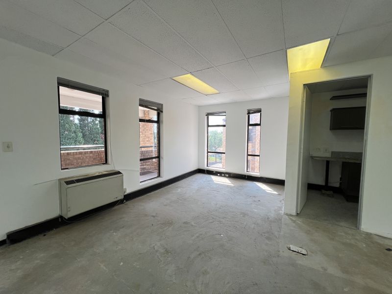 An empty office space featuring a suspended ceiling, HVAC unit, and an adjacent small kitchenette area with large windows providing natural light.