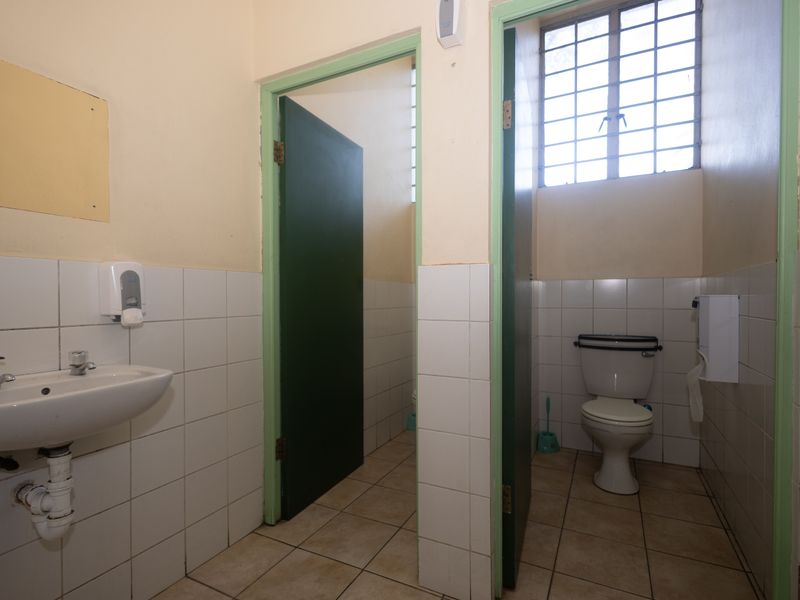 A functional commercial bathroom featuring tiled walls and floors, a wall-mounted basin, and a private toilet cubicle with a security window grille.