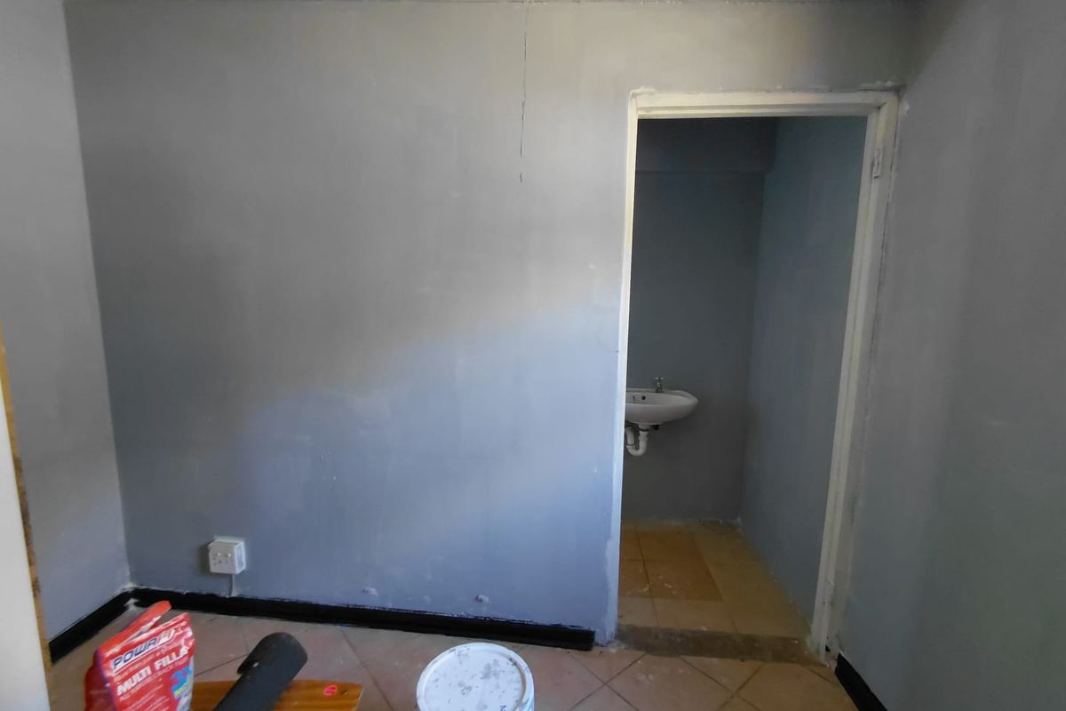 An interior view of a room under renovation, featuring grey painted walls, tiled flooring, and an adjacent small washroom with a wall-mounted basin.